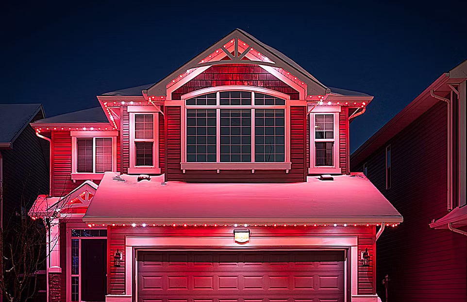 Two-story suburban house with snow on the roof, illuminated by red and white outdoor holiday lights at night.