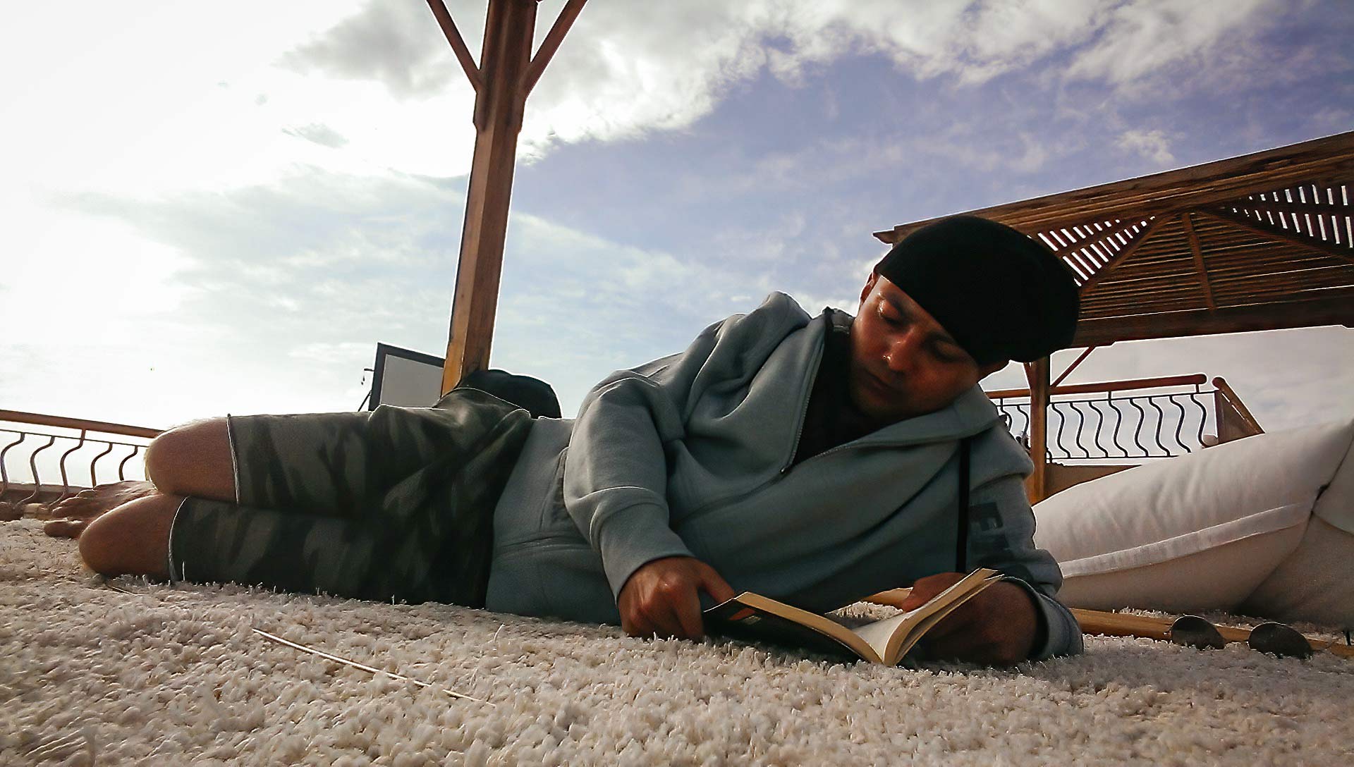 Ein Mann mit Mütze liegt auf einem Teppich auf einer Terrasse und liest in einem aufgeschlagenen Buch, über ihm bewölkter Himmel.