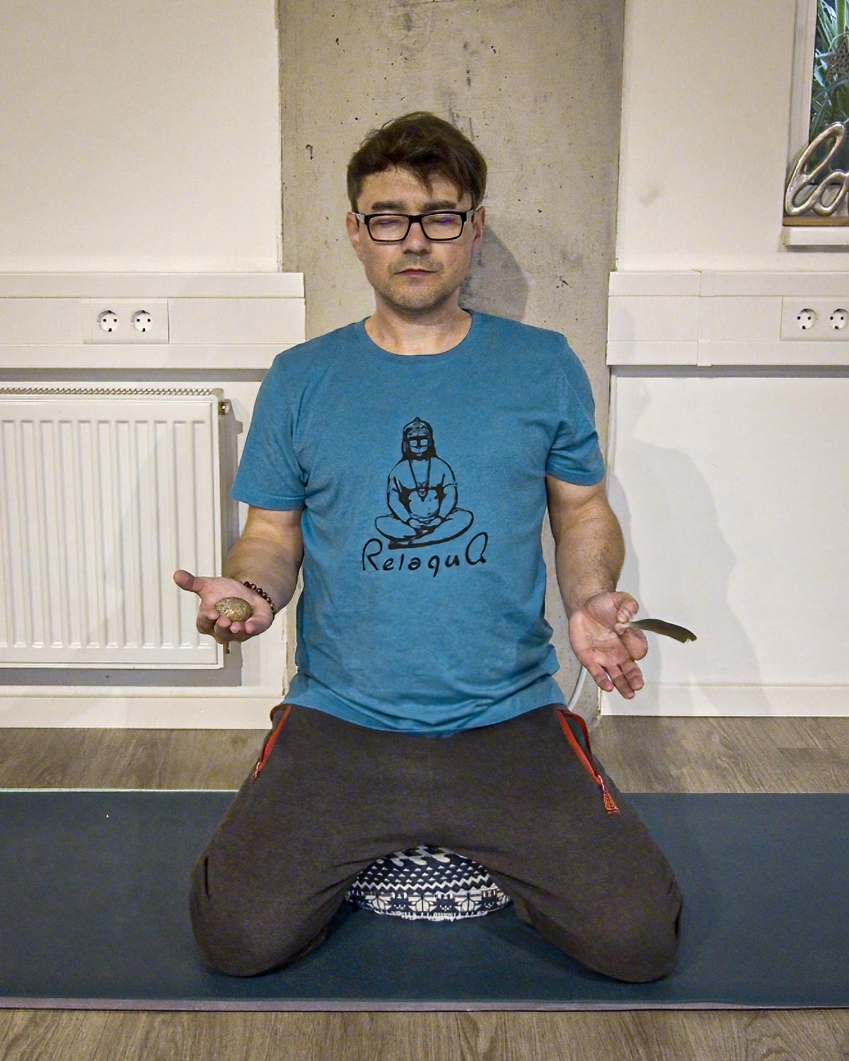 Alexander Neubert kniet mit geschlossenen Augen und geöffneten Handflächen auf einer Yogamatte im Kursraum.