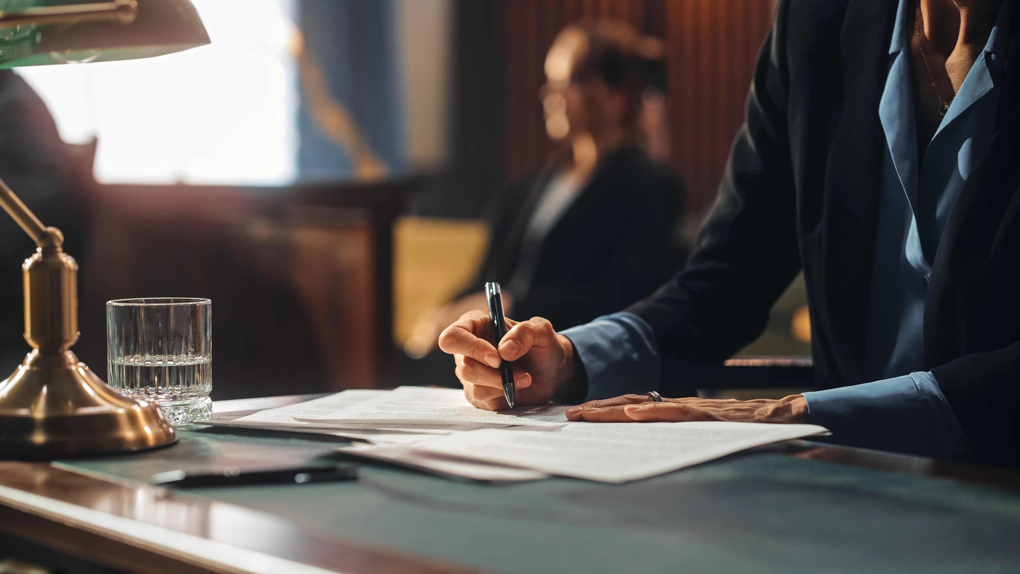 Person in business attire writing on documents at a desk with a glass of water and lamp nearby.