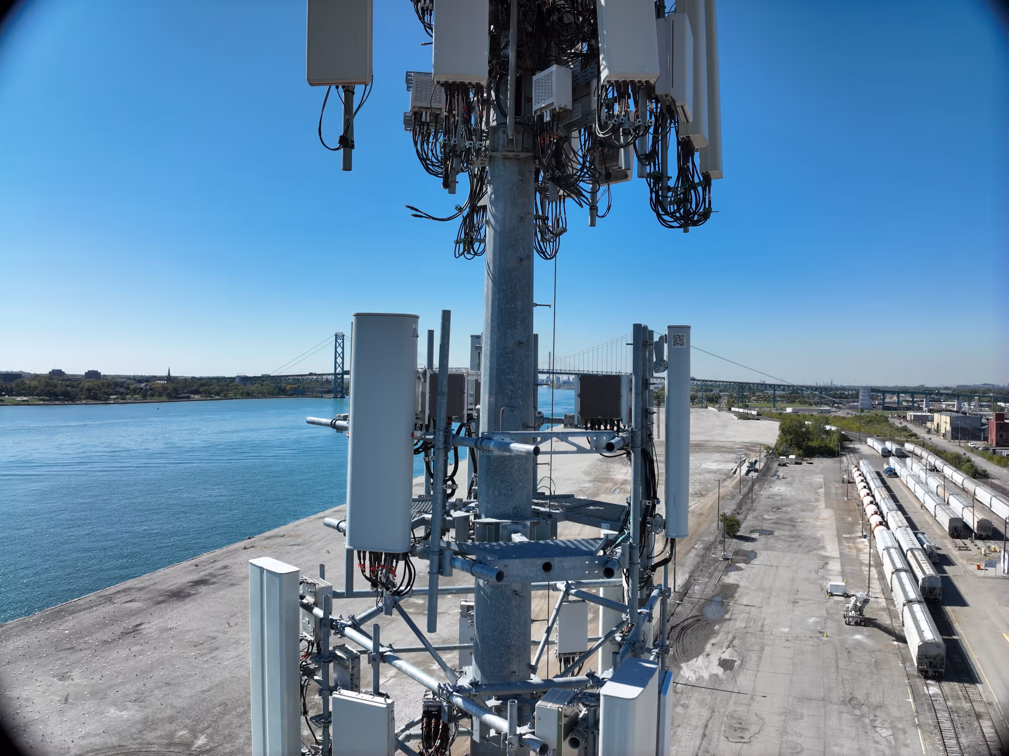 Close-up of a cellular tower with antennas overlooking a river and industrial area with train cars.