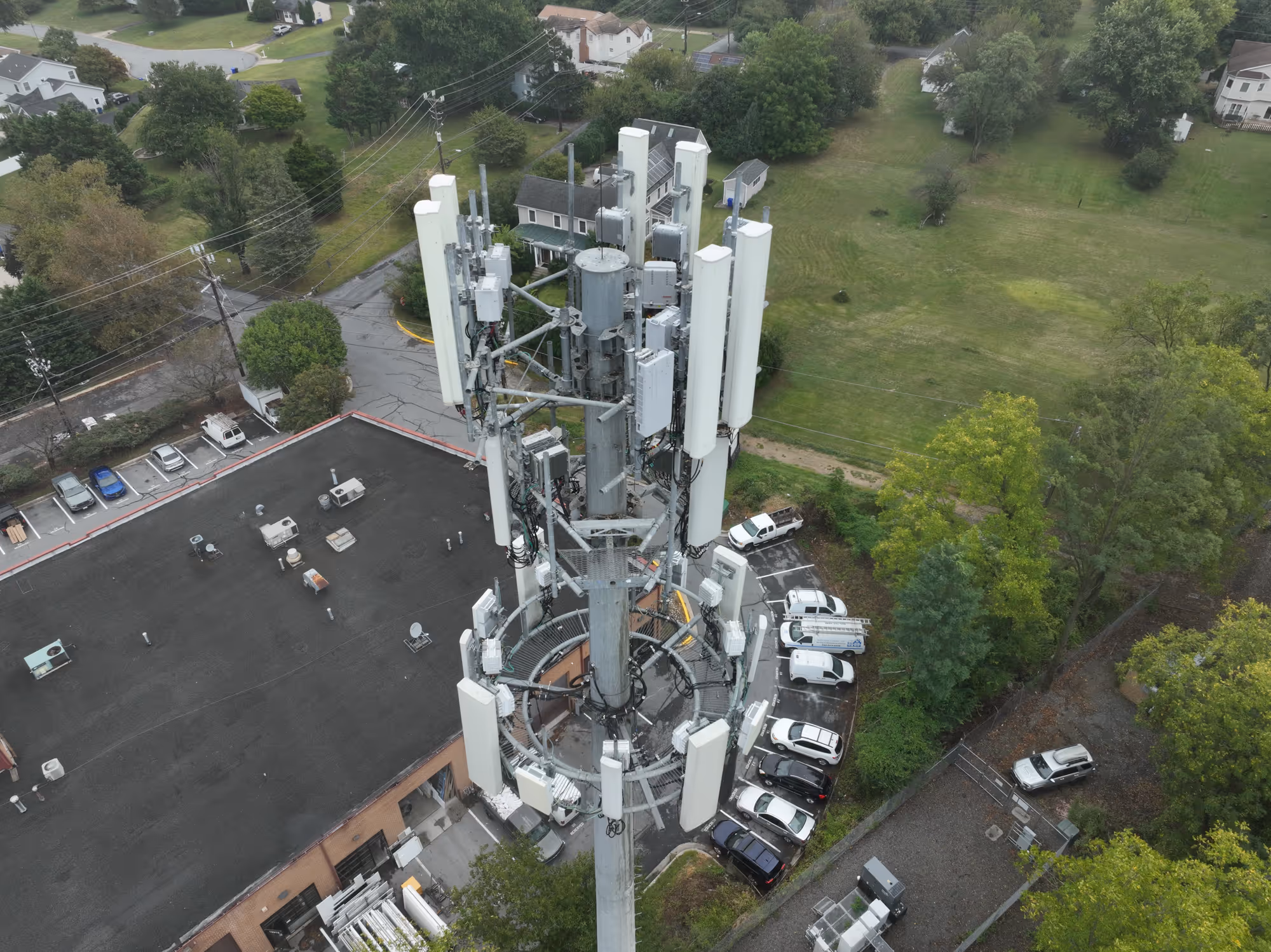 Aerial view of a large cellular tower with multiple antennas surrounded by parked cars, trees, and nearby buildings.