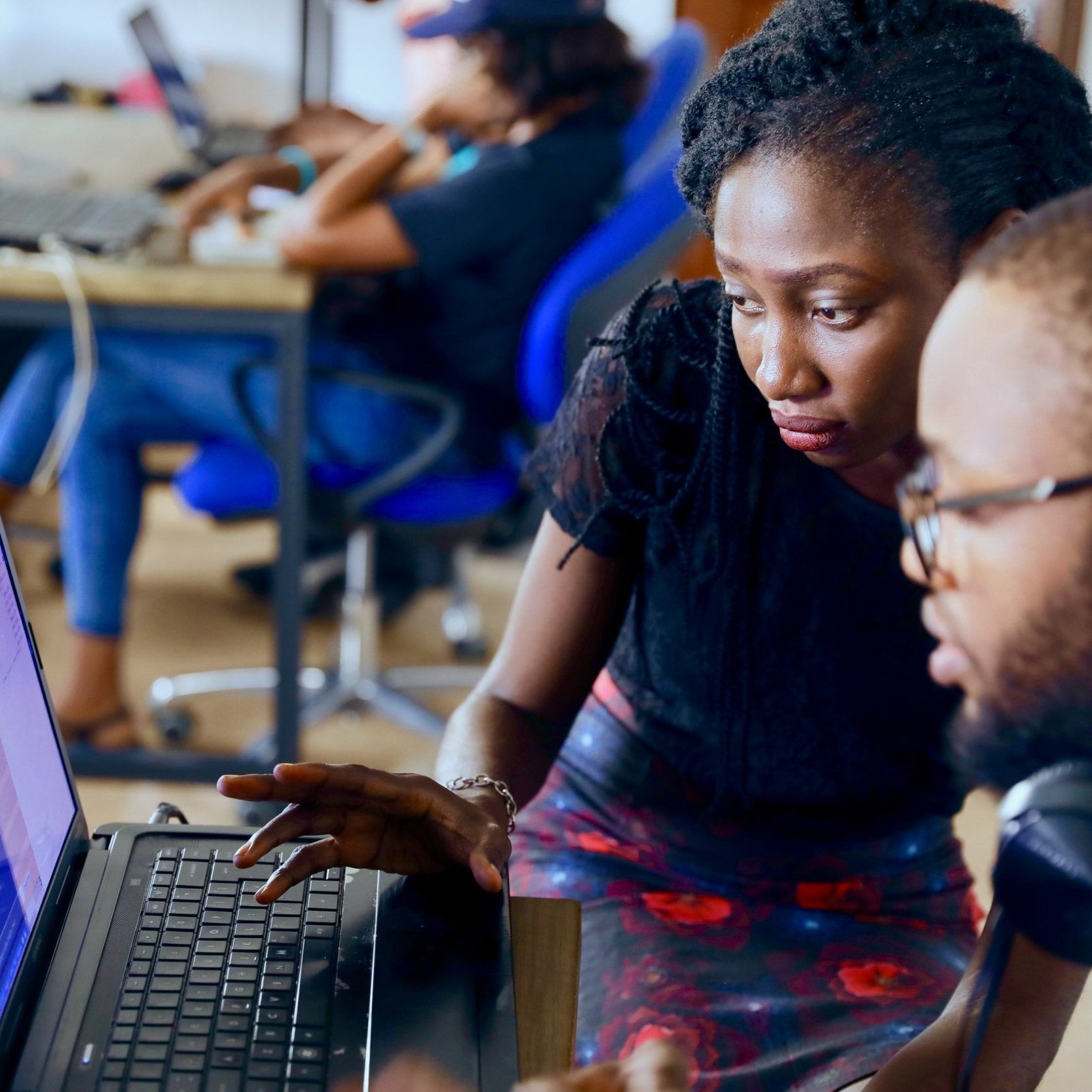 A man is sitting at his work desk looking at his laptop in front of him. A woman is stooped down next to him, helping him navigate the laptop.