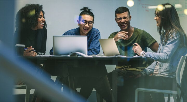 A group of four people are sitting at a table in a conference room. They are all laughing and smiling at each other. Their laptops and notebooks are in front of them on the table.