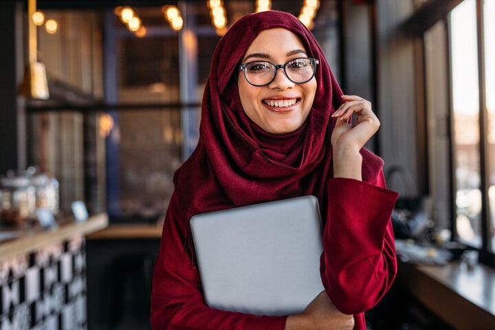 A woman standing in a coffee shop smiles at the camera. She is holding a laptop and wearing a red hijab.