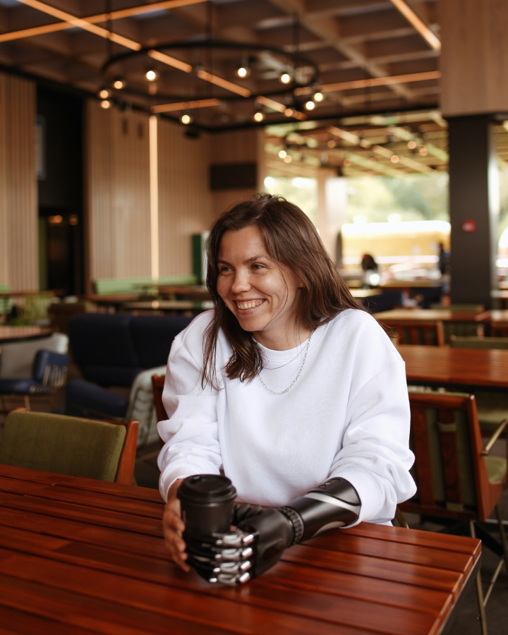 A woman sits at a table in a coffee shop. She is smiling big at something outside of the frame. She is holding a coffee in a to go cup. Her left arm is a prosthetic.