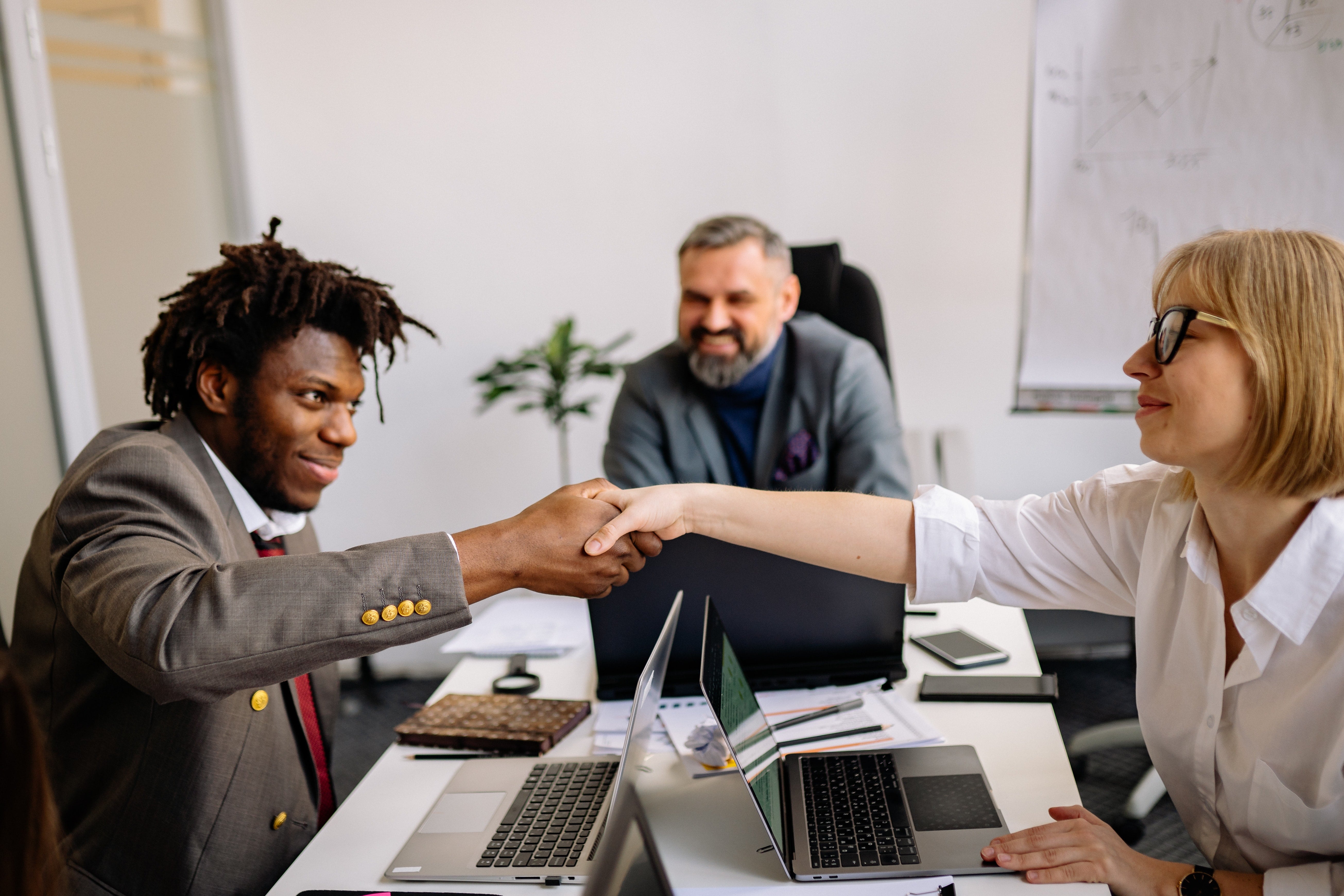 Three people sit around a desk in an office environment. Two of those people are shaking hands over the table. All three are smiling.
