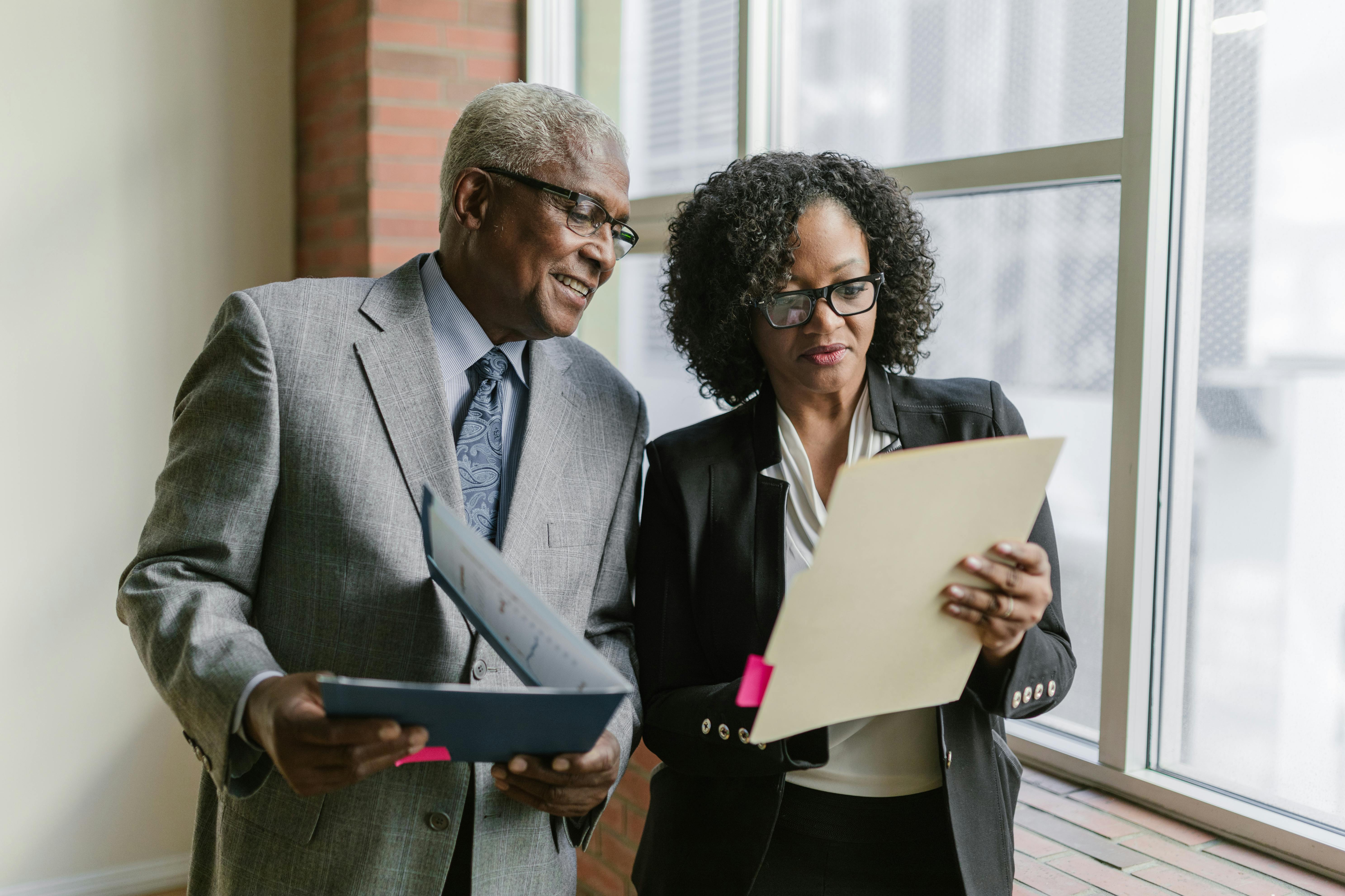 A man and a woman stand in front of a window. They are reviewing the files in their hands.