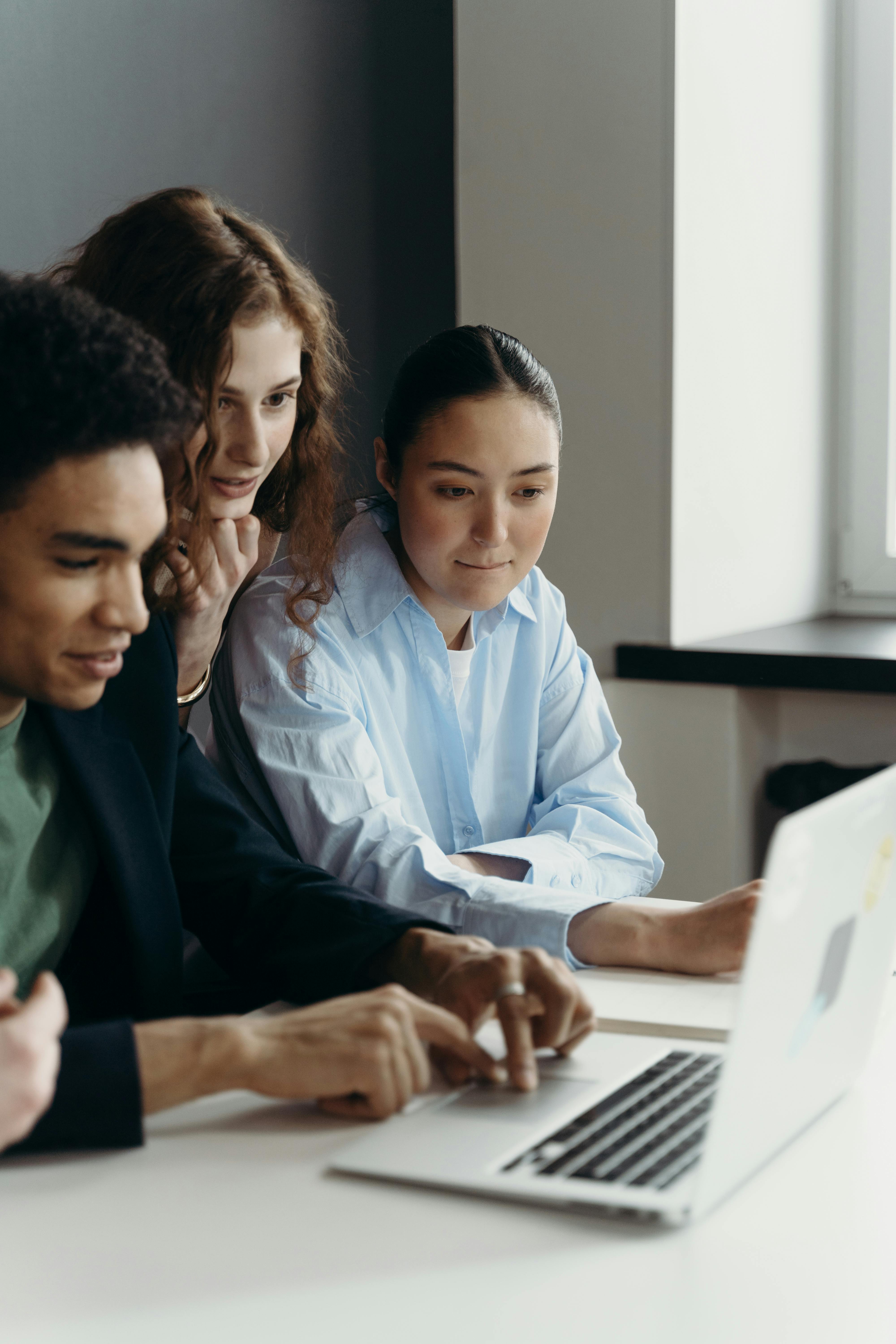 Three people surround a laptop at a table. They are looking at the screen.