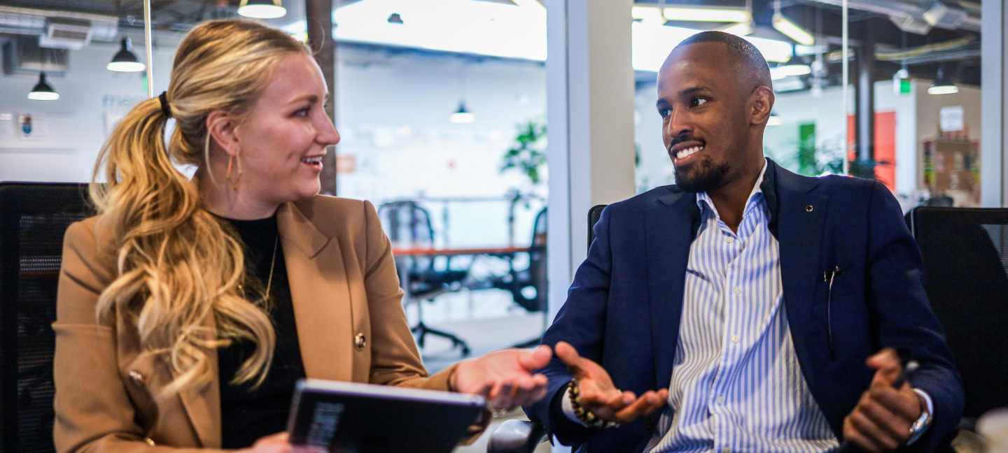 A woman sits at a table in a coffee shop. She is smiling big at something outside of the frame. She is holding a coffee in a to go cup. Her left arm is a prosthetic.