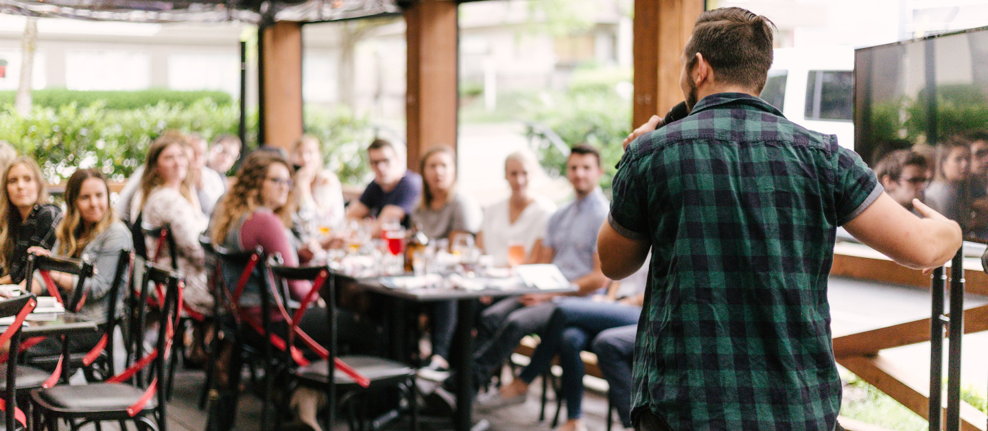 A woman sits at a table in a coffee shop. She is smiling big at something outside of the frame. She is holding a coffee in a to go cup. Her left arm is a prosthetic.