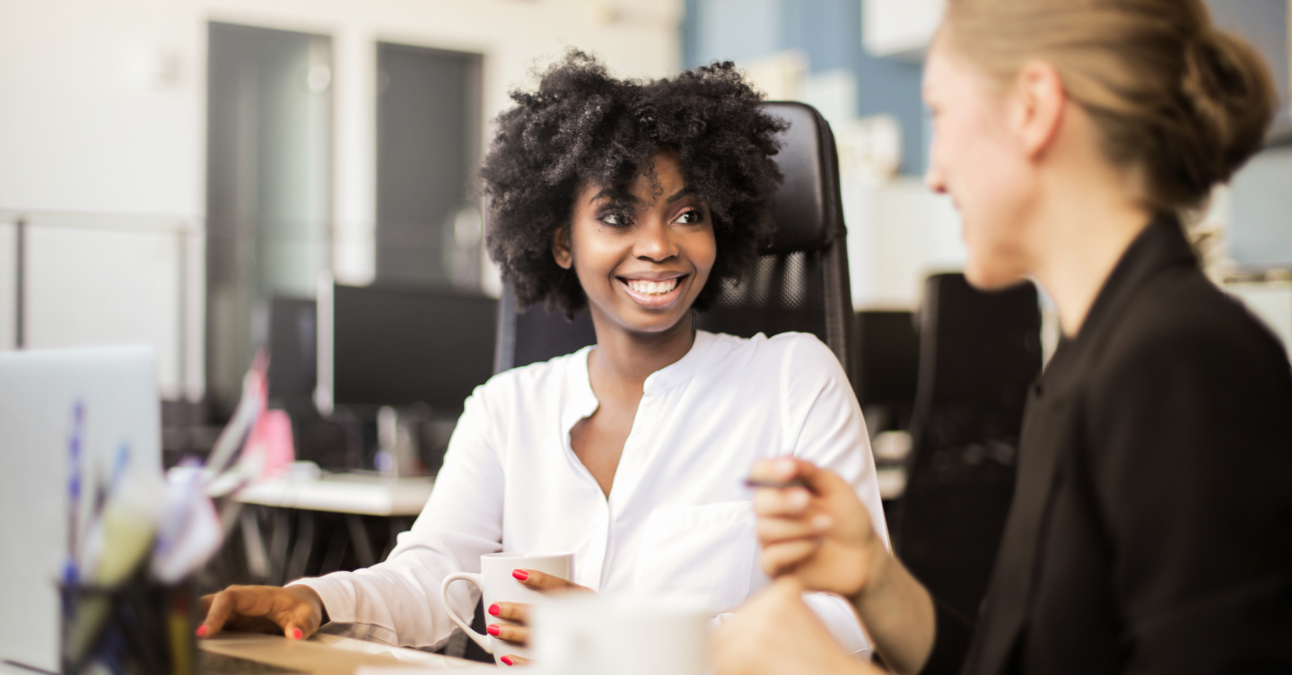 A woman sits at a table in a coffee shop. She is smiling big at something outside of the frame. She is holding a coffee in a to go cup. Her left arm is a prosthetic.