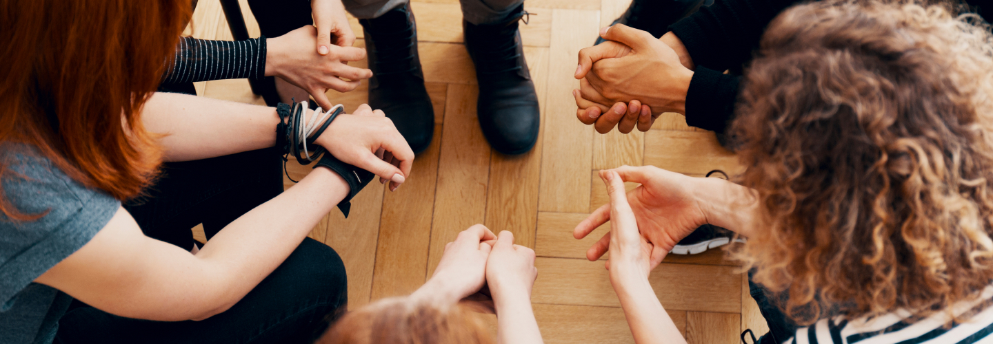 A woman sits at a table in a coffee shop. She is smiling big at something outside of the frame. She is holding a coffee in a to go cup. Her left arm is a prosthetic.