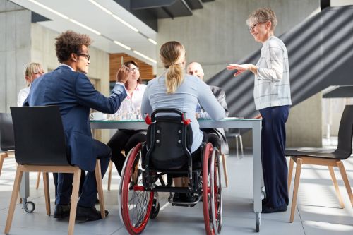 A group of people in business casual clothing are sitting at a conference table in an office. They are engaged in a positive discussion. One of the people in sitting in a wheelchair.