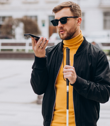 A man stands outside a building. He is holding his phone out in front of him to speak into the mic. He is wearing dark sunglasses and holding a cane in his left hand.