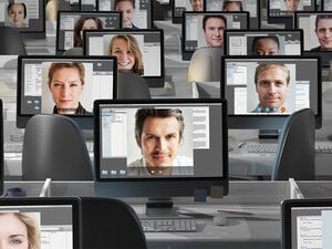 A photo of an array of computer monitors on top of desks. The desks are arranged in rows continuing out of frame. Each computer monitor has a close up image of a different person's face.