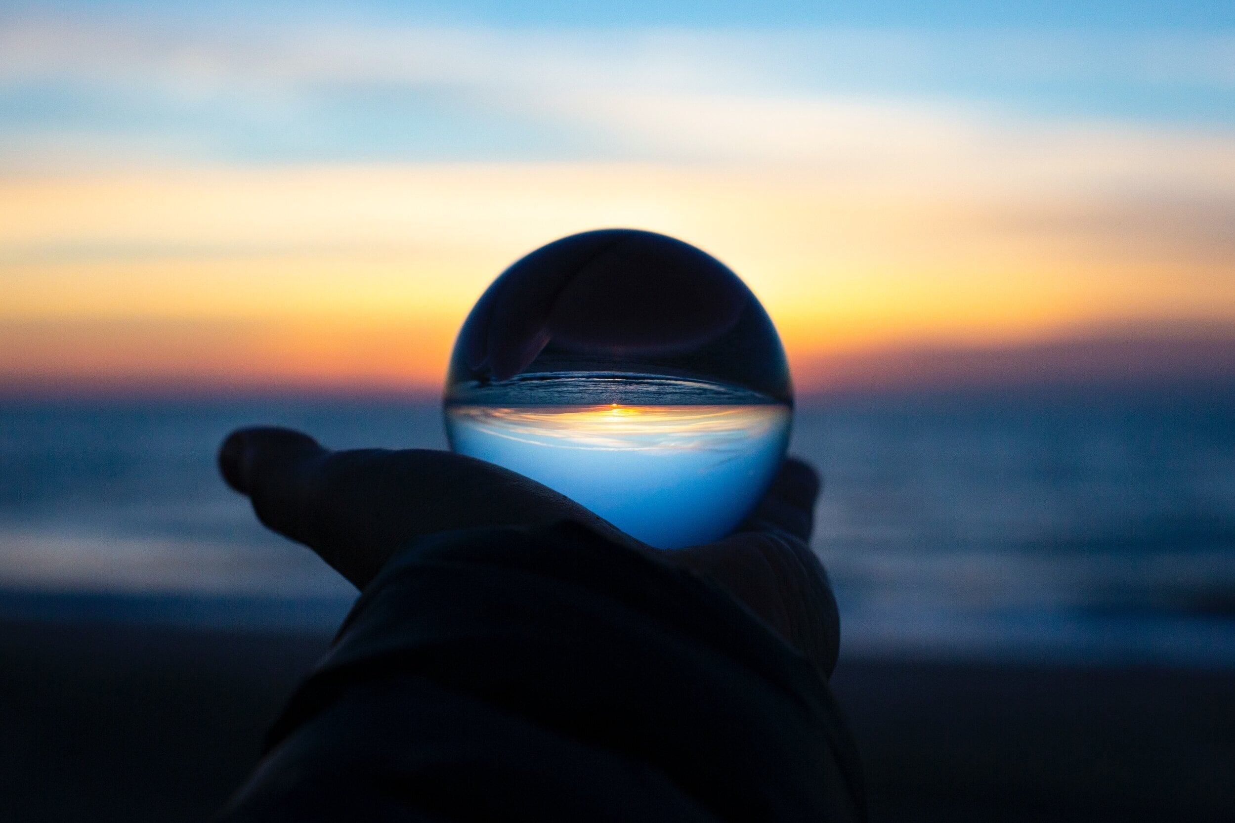 A closeup of a hand holding a glass orb. The orb is reflecting the sunset over the ocean in front of it.