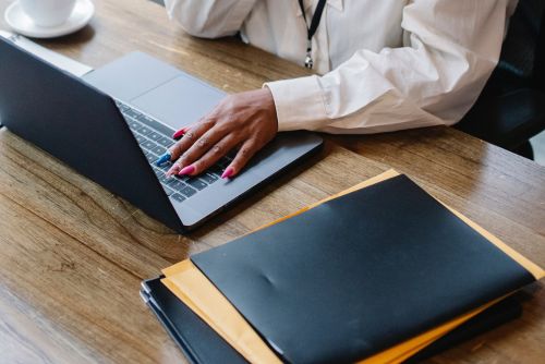 A close up image of a working desk. There are files on the desk and a laptop that a woman is typing on.