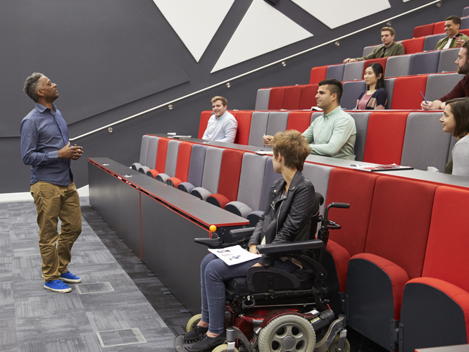 A photo of a professor speaking at the front of a classroom. There are several students seated, including one person sitting in a wheelchair.