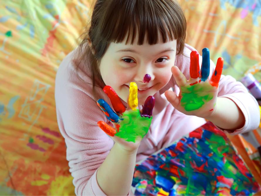 A photo of a girl with Down Syndrome holding up her hands to the camera. They are covered in paint as she has been fingerpainting on a canvas. 