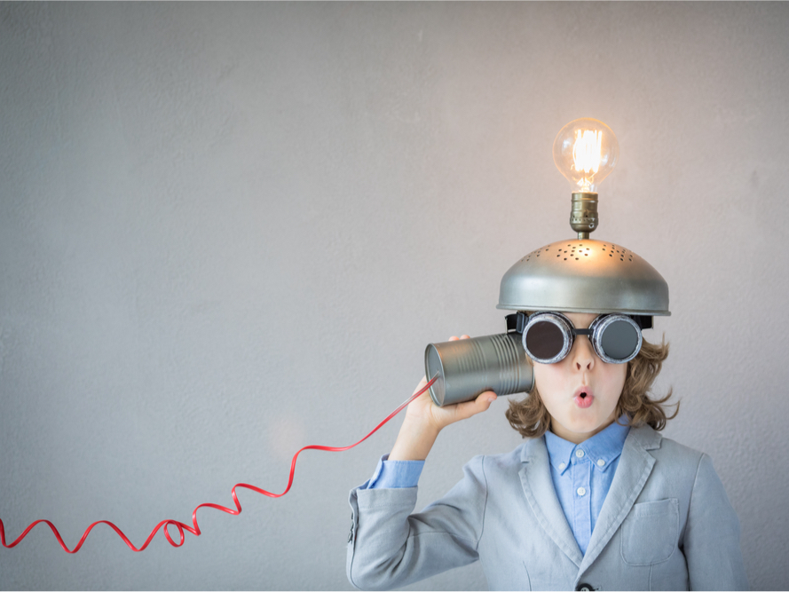A photo of a kid wearing a metal hat with a lightbulb on top and goggles. They're holding an empty can to their ear which is connected to a wire.