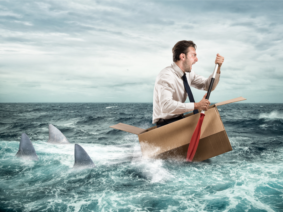 An image of a man sitting in a cardboard box in the middle of the ocean. He is furiously paddling away from the shark fins behind him.