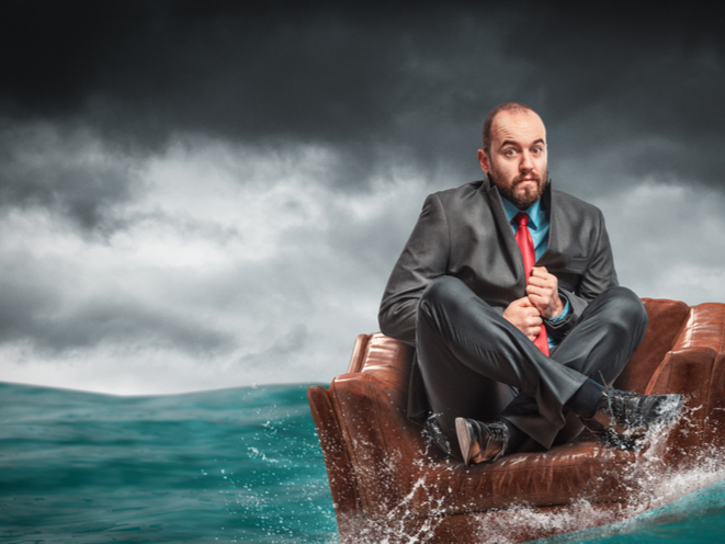 An image of a man sitting on a chair in the middle of the ocean. He has an anxious look on his face.