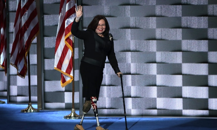 Asian woman with double amputation (Tammy Duckworth) smiles and walks across the stage on two prosthetic legs and carries a cane.