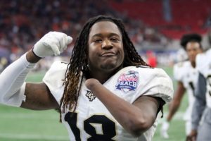 African American man with dreadlocks wearing a football; uniform and pads holds up his arms. You can see that his left arm is missing a hand below the wrist.