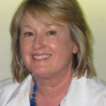 Smiling woman with blond hair wearing a white Blazer and a Blue beaded necklace.