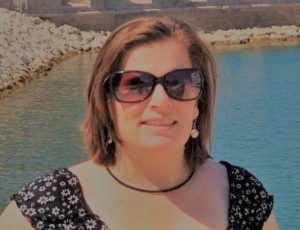 Long Description Image of a smiling brown haired woman wearing sunglasses and standing in front of a body of water.