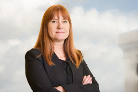 A confident red-headed woman in a black suit stares at the camera as if it is a mere hurdle to be conquered.