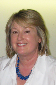 Smiling woman with blond hair wearing a white Blazer and a Blue beaded necklace.