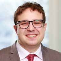 Image of a smiling young caucasian man with brown hair who is wearing glasses a a brown jacket, white shirt and red tie,