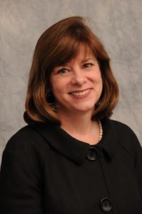 A smiling woman with brown hair wearing a black top with pearls.