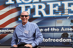 Image of a smiling man in a blue shirt and sunglasses sitting in a wheelchair in front of a bus on the side which the word Freedom is visible.