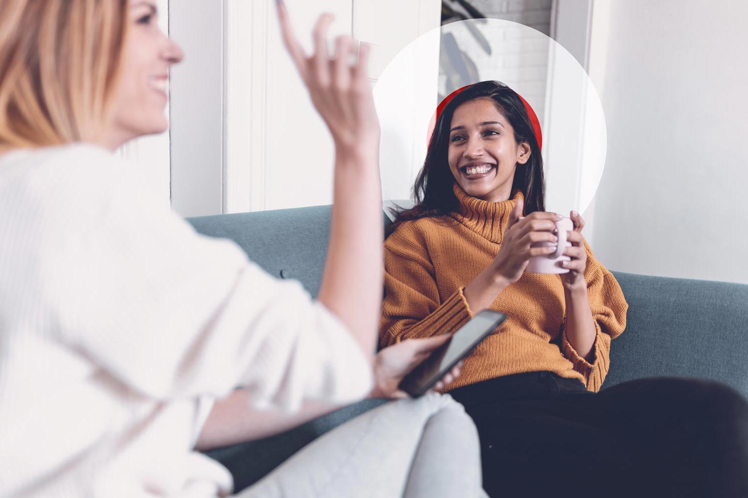 A young woman with long dark hair, wearing a mustard yellow turtleneck sweater, sits on a blue couch smiling broadly while holding a mug. She is listening to another woman in the foreground who is gesturing and smiling while holding a phone. A faint, semi-transparent white circle graphic is centred behind the seated woman’s head.