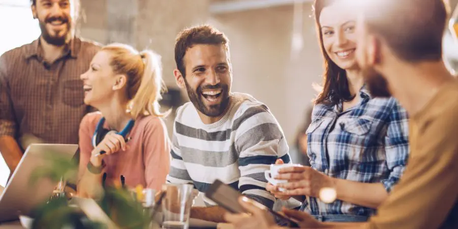 Group of joyful colleagues sharing a light moment over coffee in a sunny office environment.