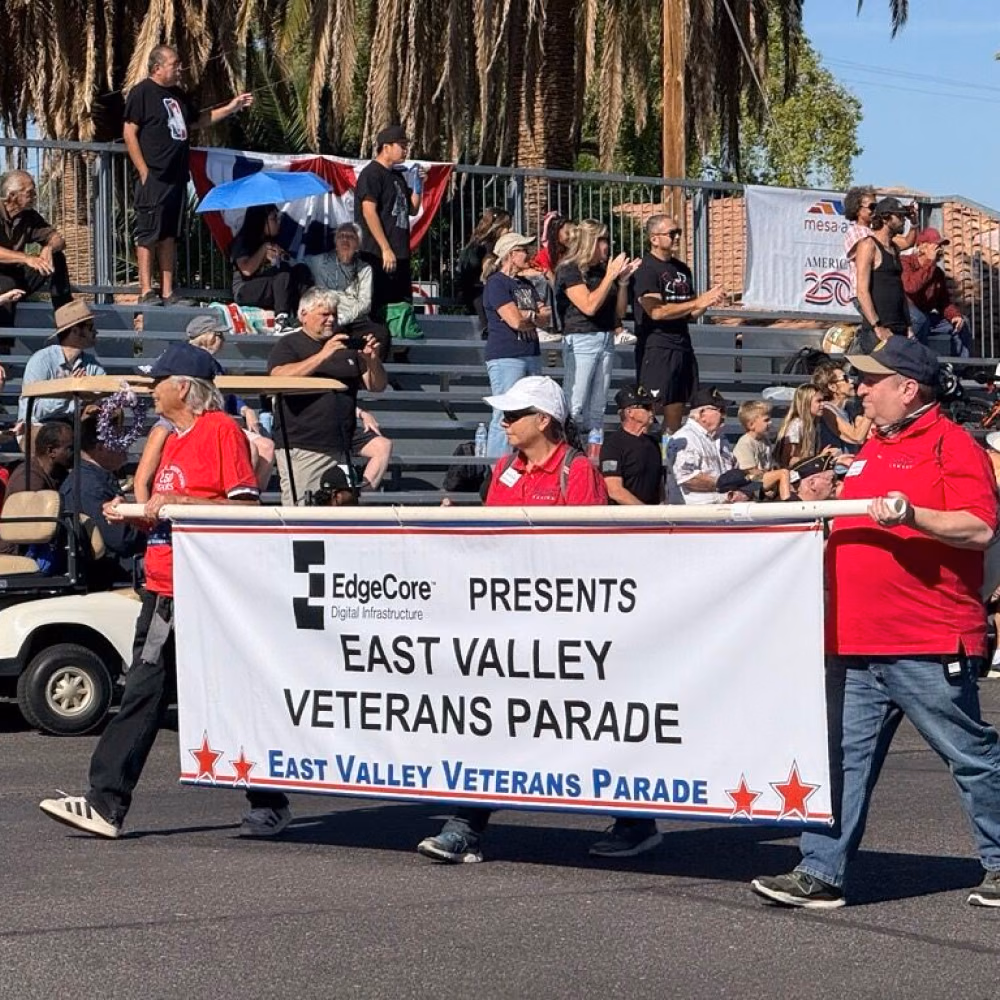 Three people in red shirts holding a banner for the East Valley Veterans Parade with spectators behind bleachers.