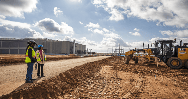 Two construction workers in safety vests and helmets standing by a dirt road at a construction site with a large CAT motor grader leveling the ground on the right and industrial buildings in the background under a partly cloudy sky.