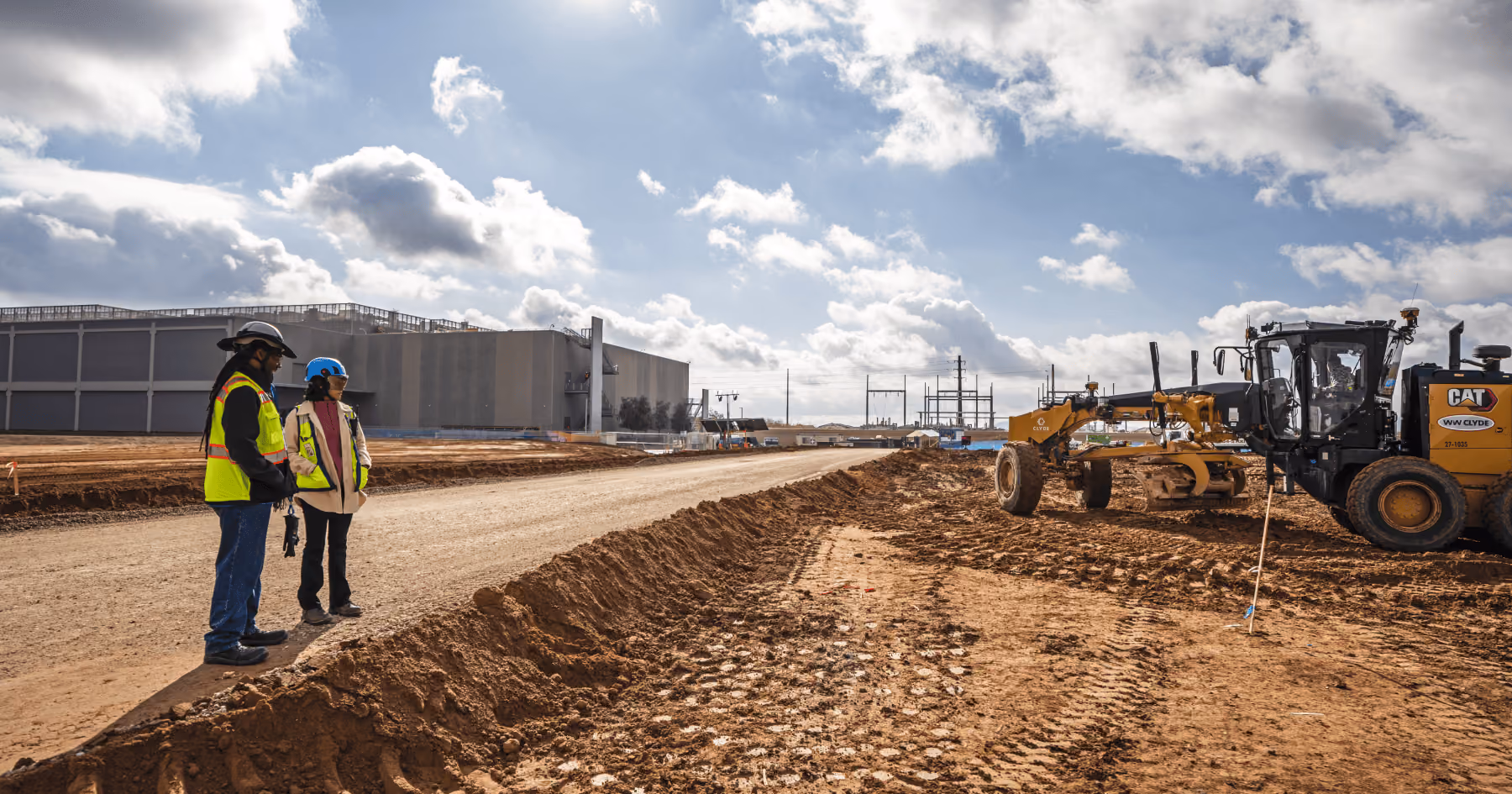 Two construction workers in safety vests and helmets standing by a dirt road at a construction site with a large CAT motor grader leveling the ground on the right and industrial buildings in the background under a partly cloudy sky.