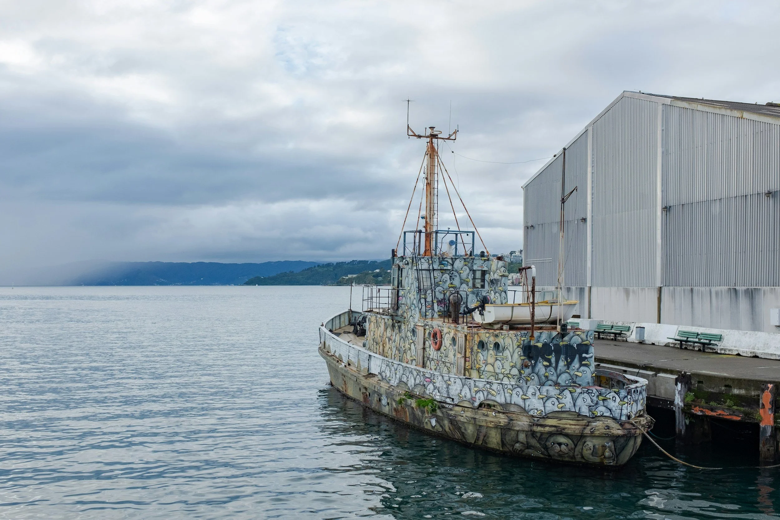 Rusty boat docked near a grey warehouse, covered in cartoonish graffiti of birds and animals on a cloudy day.