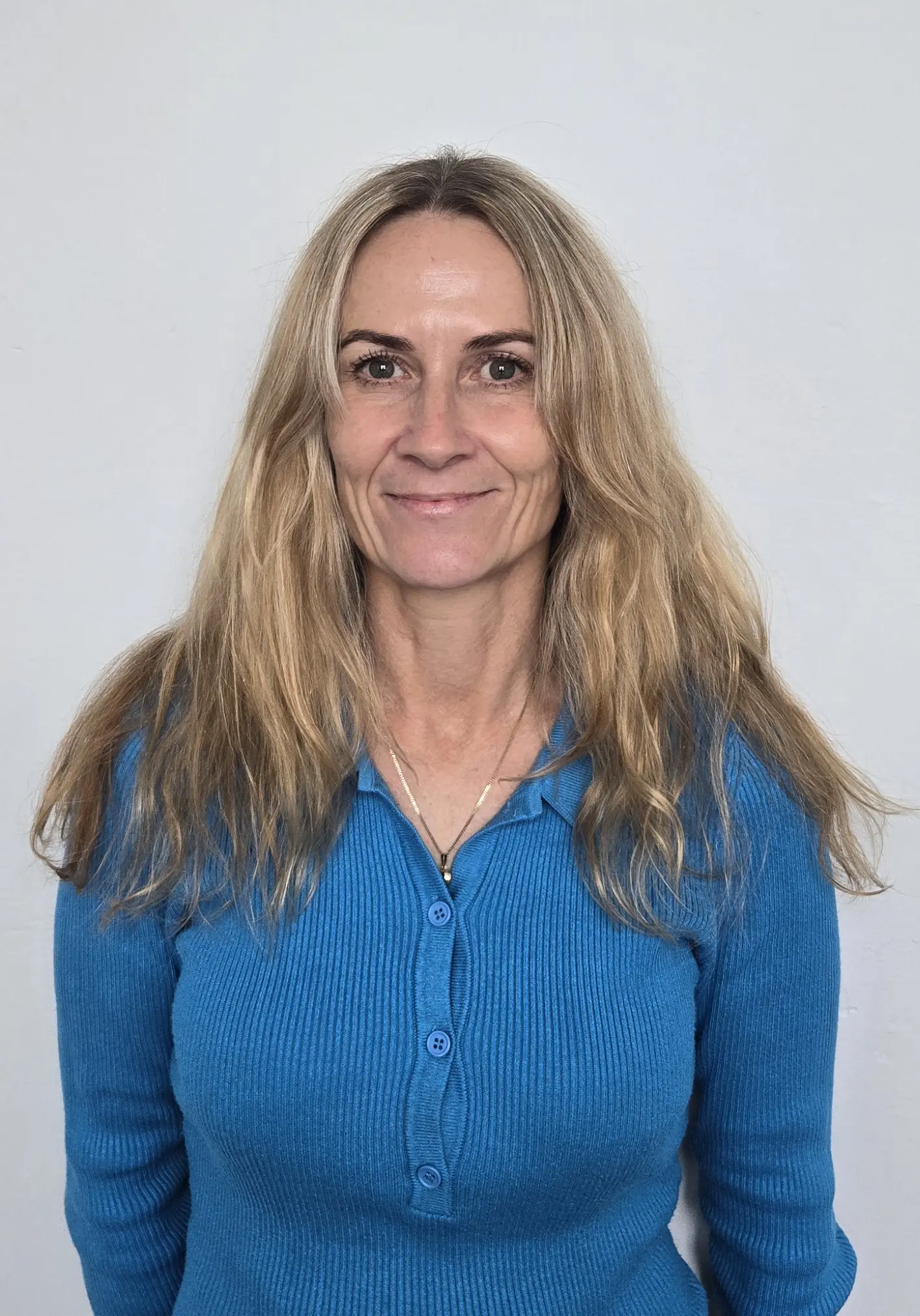 Smiling woman with long blonde hair wearing a blue ribbed buttoned shirt against a light grey background.