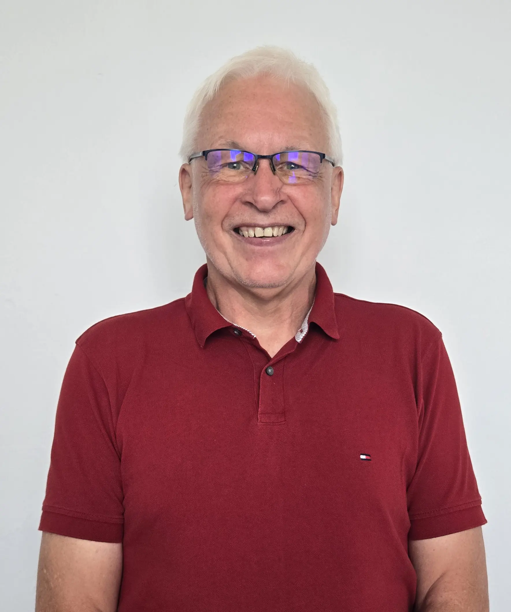 Smiling elderly man with white hair wearing glasses and a red polo shirt against a plain light background.