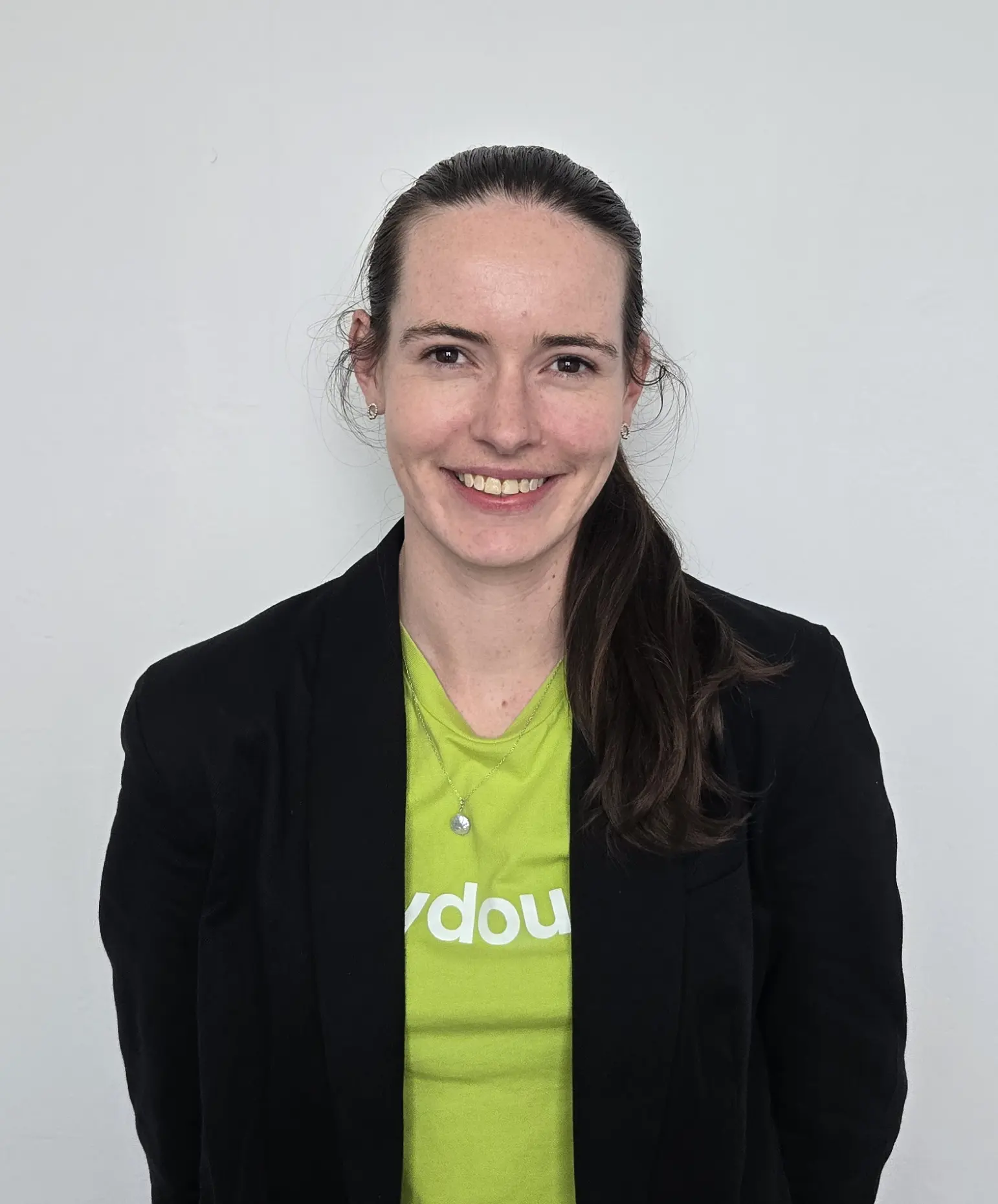 Smiling woman with long dark hair tied back, wearing a black blazer over a green shirt with partially visible white text, and a silver necklace.