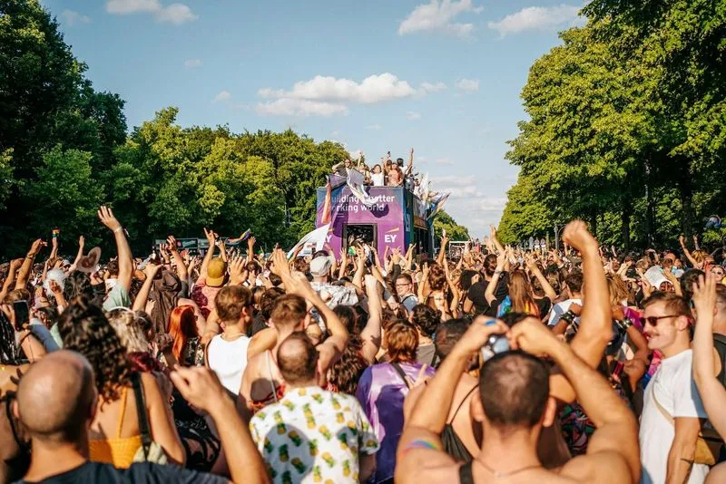 A lively crowd celebrates at an outdoor parade on a sunny day, with people raising their hands and a colorful float in the background under blue skies.