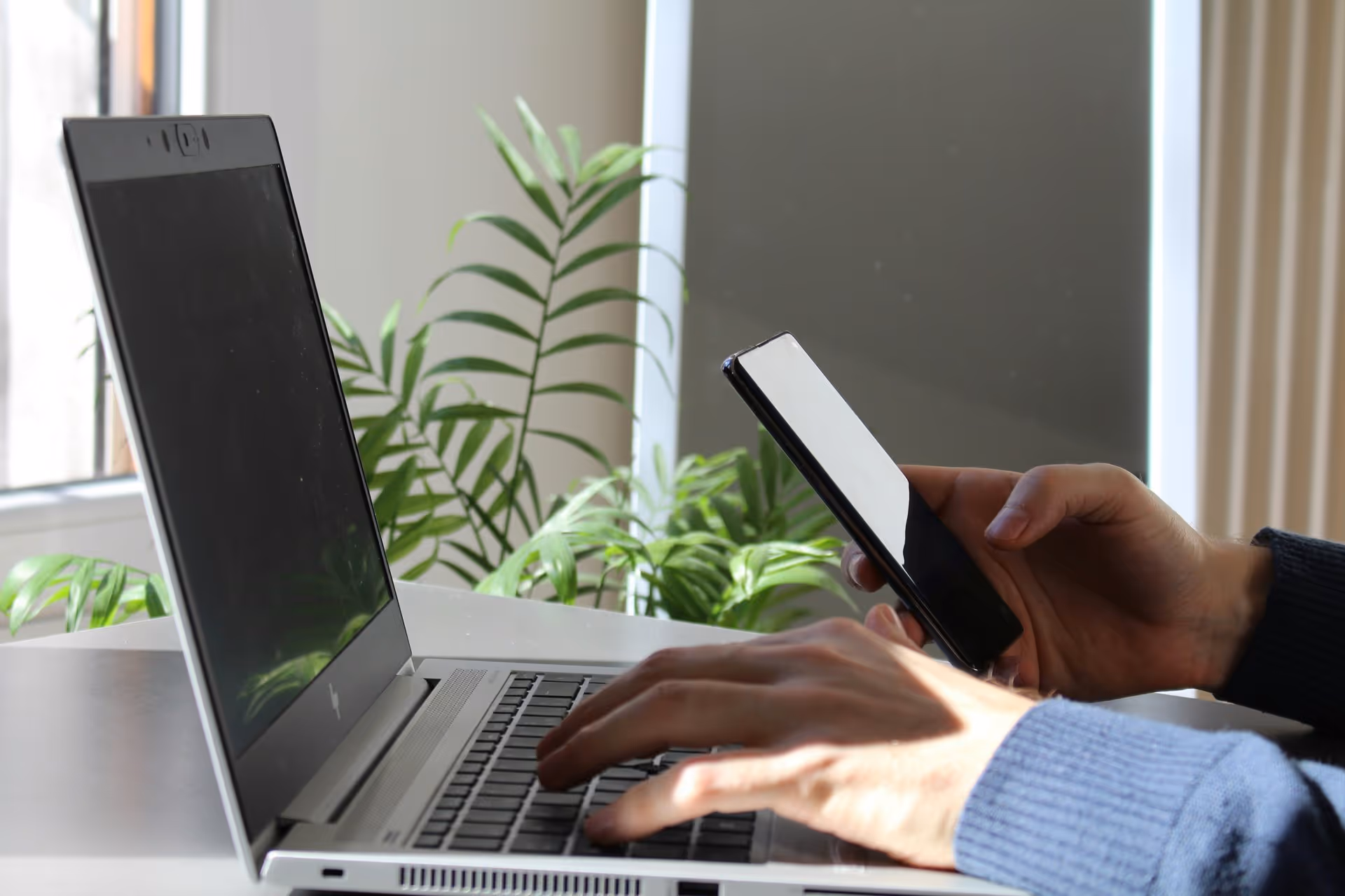 Hands holding a smartphone and typing on a laptop in a brightly lit room with green plants. The scene conveys multitasking or technology use.