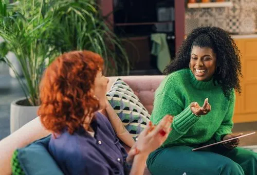Two women sit on a sofa in a cozy room, engaged in a lively conversation. One wears a green sweater, the other a blue shirt. Both are smiling warmly.