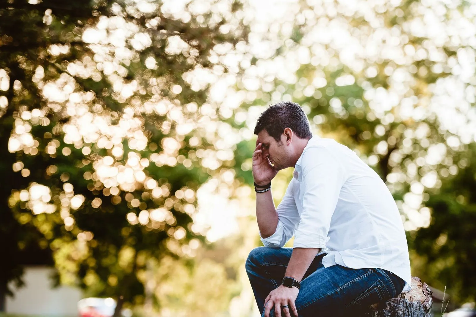Man in white shirt and jeans sits on a tree stump in a park, covering his face with one hand, appearing stressed. Sunlight filters through leafy trees.