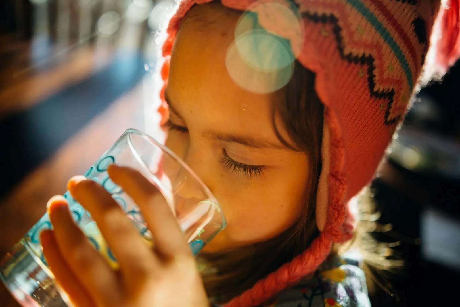 A child in a pink knit hat drinks from a glass of water, eyes closed, absorbing warmth from sunlight streaming in. The mood is peaceful and cozy.